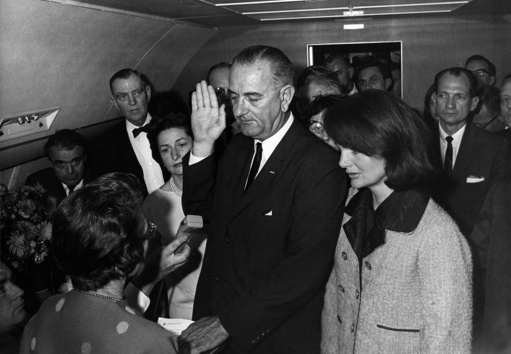 Lyndon B. Johnson taking the oath of office aboard Air Force One  two hours and eight minutes after the assassination of John F. Kennedy, Dallas, Texas. Jackie Kennedy (right), still in her blood-soaked clothes, looks on. 
